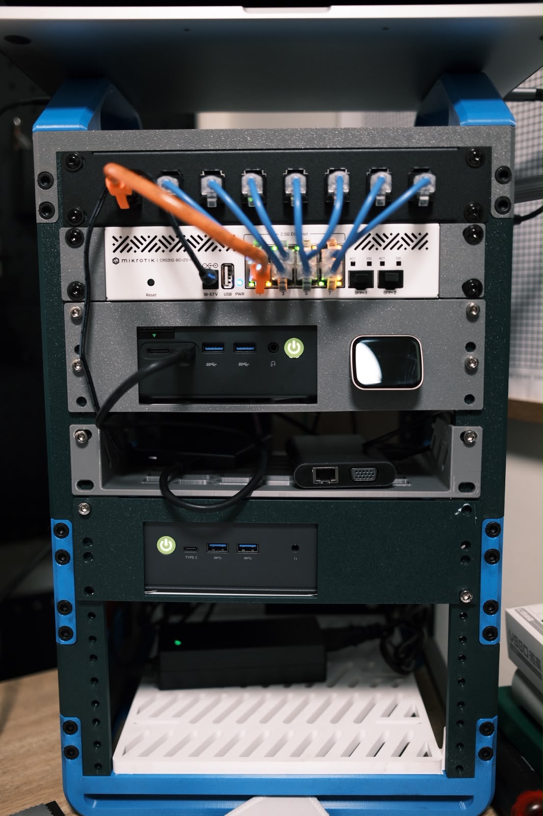 A 10-inch, 3D-printed server rack holding a MikroTik CRS310 switch, a JetKVM, and two GMKtec mini PCs, patched together with blue and orange cables